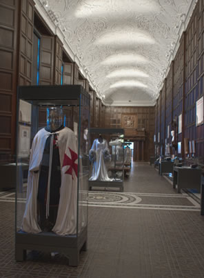 The exhibit hall of the Folger, with a concave, scrolled plaster ceiling and a tile floor, display cases in the middle with a knights costum in the foreground. 